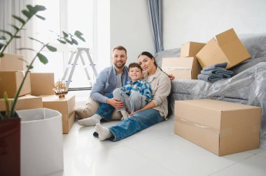 Üç kişilik mutlu bir aile yeni dairelerinde oturmuş kutuları taşıyarak, taşınmayı kutluyorlar.