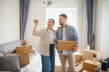 Mutlu çift ev sahipliğini kutluyor, ellerinde ev anahtarları ve karton kutular yeni evlerinin oturma odasında.
