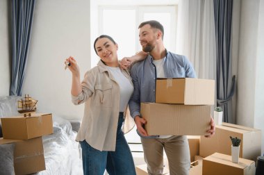 Genç çift yeni eve taşınmayı kutluyorlar. Kadın anahtarları tutarken erkek içeride kutuları taşıyor.