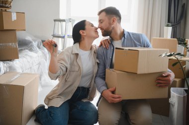 Anahtarlarla poz veren mutlu genç çift, taşınma sırasında karton kutularla çevrili yeni bir ev satın almalarını kutluyorlar.