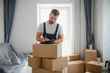 Üniformalı bir adam, taşınma servisi sırasında karton kutularla çevrili bir pano ve kalemle çalışıyor.