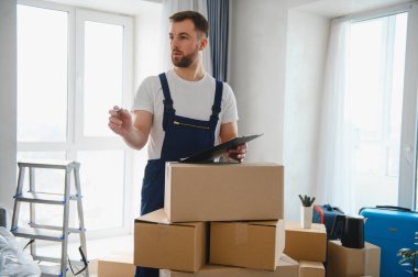 Üniformalı bir adam elinde bir pano, yeni bir dairede karton kutuların arasında duruyor, teslimatı garantiliyor ya da paketleniyor.