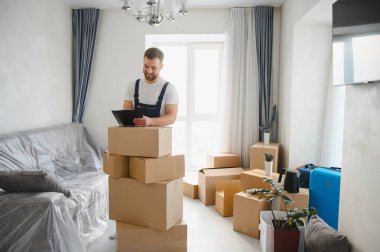 Profesyonel nakliye şirketi çalışanı tulum giyiyor, panoyu kontrol ediyor, yeni bir evde karton kutuları istifliyor.