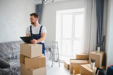 Profesyonel bir nakliyeci elinde bir pano, bir ev taşınması sırasında karton kutuların arasında duruyor, sorunsuz bir teslimatı garantiliyor.