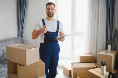 Üniformalı bir adam elinde bir pano, bir odada duruyor ve karton kutularla taşınmaya hazırlanıyor.