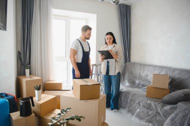 Genç bayan müşteri ve profesyonel nakliyeci eşyaları paketlenmiş kutularla yeni bir daireye yerleştirirken envanteri kontrol ediyorlar.