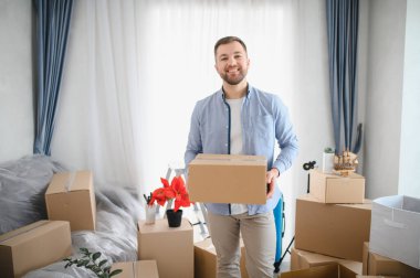 Gülümseyen adam parlak bir odada duruyor, karton bir kutu taşıyor, taşınma sırasında diğer paketlenmiş kartonlarla çevrili.
