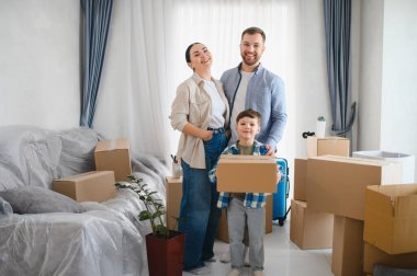 Yeni dairelerinde mutlu bir aile duruyor, taşınma gününü kutluyorlar karton kutular ve taşınma için paketlenmiş mobilyalarla.