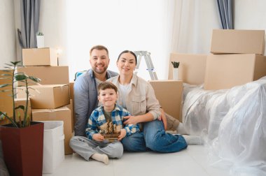 Karton kutularla çevrili yerde oturan gülümseyen aile taşınma günlerini ve evlerinde yeni başlangıçları kutluyorlar.