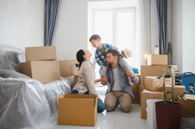 Babasının omuzlarında çocuğu olan aile yeni evinde gülümsüyor karton kutularla çevrili, taşınmayı kutluyorlar.