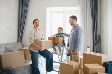 Mutlu aile yeni dairede karton kutuların arasında duruyor, ebeveynler ve çocuklar taşınıyor veya taşınıyor.