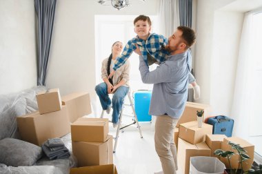 Genç bir aile boş bir odada yeni bir başlangıcı kutluyor. Kutularla çevrili. Heyecan ve yeni bir başlangıç anlamına geliyor.