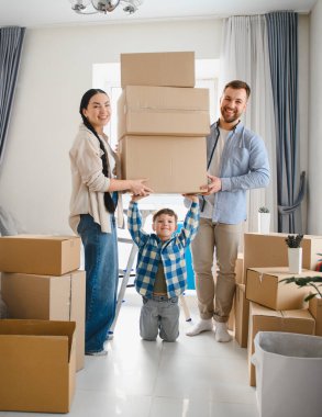 Neşeli ebeveynler ve çocuklar yeni bir daireye taşınıyorlar. Hepsi sırıtıyorlar ve kutuları taşıyorlar.