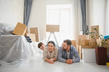 Ebeveynler ve çocuklar yeni başlangıçlarından dolayı mutlu ve heyecanlı bir şekilde kutuları taşıyarak yerde dinleniyorlar.