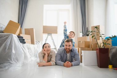 Genç bir aile, anne-baba ve oğul yerde yatıp yeni evlerine taşınmayı kutluyorlar.