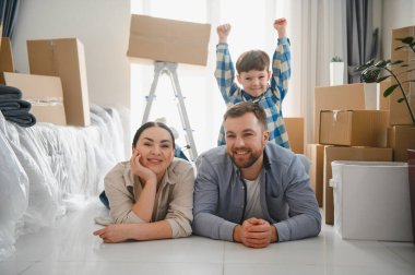 Genç aileler ve heyecanlı çocuklar yeni evlerinin zemininde karton kutularla çevrili yatıyorlar, taşınma gününü kutluyorlar.