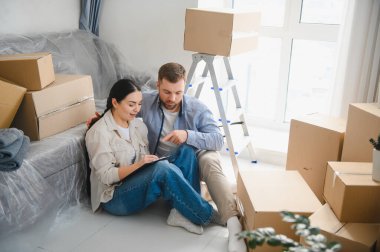 Genç bir çift yerde oturmuş belgelere bakıyor ve yeni dairede karton kutularla çevrili planlarını tartışıyorlar.