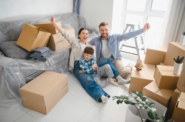 Neşeli ebeveynler ve çocuklar, kutuları taşıyarak yerde oturuyorlar, yeni bir eve taşınmayı kutluyorlar.