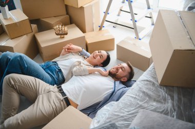 Gülümseyen genç bir çift, yeni dairelerinde taşınan kutular ve mobilyalarla birlikte yerde yatıyorlar.