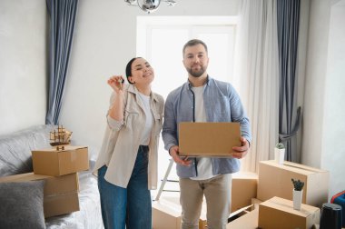 Yeni ev anahtarları ve taşınma kutusu taşıyan gülümseyen bir çift, yeni boş dairelerinde karton kutularla çevrili.