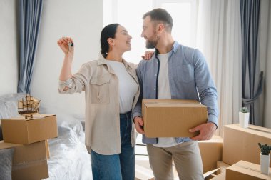 Genç çift taşınma gününü kutluyor, yeni ev anahtarlarını ve taşınma kutusunu tutuyor, yeni evlerinde neşe ve mülkiyet hissediyorlar.