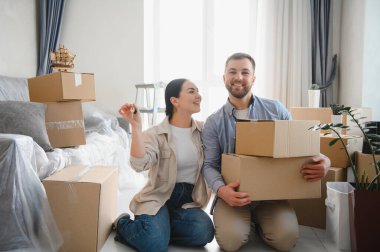 Genç çift yeni bir eve taşınmayı kutluyorlar. Karton kutularla çevrili yerde oturuyorlar ve ellerinde evin anahtarları var.
