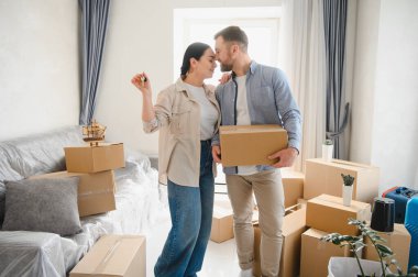 Genç çift yeni evlerine taşınmayı kutluyorlar, ellerinde bir ev anahtarı ve karton kutu var, etrafları eşyalarla çevrili.