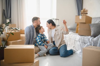 Karton kutuların arasında oturan mutlu bir aile, yeni evlerini kutluyor ve ev anahtarlarını tutuyor, taşınma kavramını