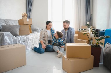 Genç bir aile, aileler ve çocuklar, yeni evlerinde paketlenmiş karton kutularla birlikte yerde dinleniyorlar.