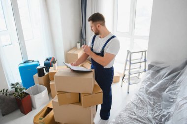 Profesyonel nakliyeci karton kutular ve mobilyalarla çevrili bir pano kontrol ediyor.