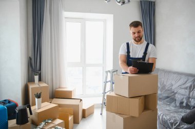 Profesyonel nakliyeci boş bir odada duruyor, karton kutuların yanındaki panoya bakıyor. Taşınma günü konsepti