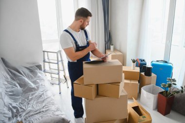 Profesyonel bir nakliyeci boş bir odada duruyor, yeni eve taşınma servisi için karton kutuların yanındaki panoyu kontrol ediyor.