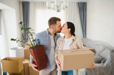 Mutlu çift öpüşüyor, ev bitkisi ve karton kutu tutuyor, yeni dairenin taşınmasını kutluyorlar.