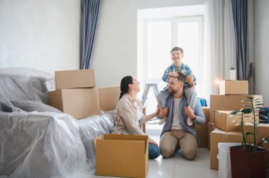 Genç bir aile, anne, baba ve oğul, mutlu bir şekilde yeni evlerine taşınıyorlar, etrafı karton kutularla çevrili.