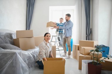 Mutlu bir aile yeni bir eve taşınıyor, karton kutuları açıyor ve parlak, ferah oturma odalarında gerekli şeyleri düzenliyor.