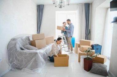 Genç bir aile yeni bir daireye taşınıyor, aileler ve çocuklar karton kutulardan eşyaları çıkarmada işbirliği yapıyorlar.