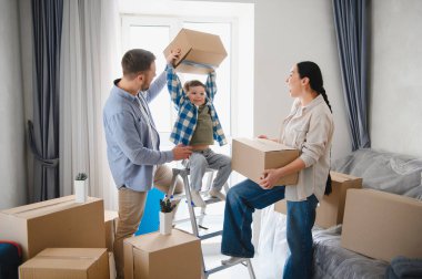 Mutlu aile yeni bir dairede karton kutuları açıyor, ebeveynler ve çocuklar eşyalarını düzenlemek için işbirliği yapıyorlar.