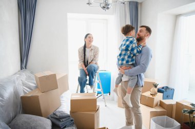 Mutlu bir aile yeni evlerinde eşyalarını boşaltıyor, baba oğlunu kucağında tutuyor, anne ise etrafı kutularla çevrili bir merdivende oturuyor.