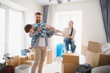 Genç bir aile taşınmayı kutluyor, bir baba oğlunu kaldırırken anne izliyor, karton kutularla çevrili.