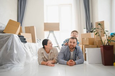 Çocuklu bir aile yerde yatıyor, gülümsüyor ve yeni evlerinde kutuları taşıyarak anın tadını çıkarıyor.