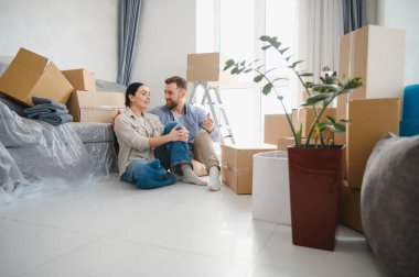 Yeni evlerinin zemininde oturan gülümseyen çift taşınma sırasında karton kutularla çevrili olarak yeni bir hayata başlıyorlar.