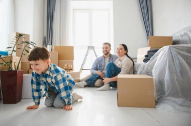 Genç bir aile taşınıyor, oğlu yeni bir dairede sürünüyor, ailesi arkada oturuyor, etrafı karton kutularla çevrili.