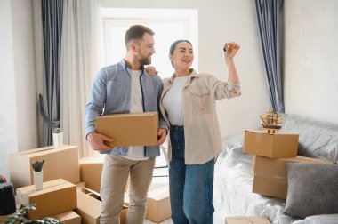 Mutlu çift ellerinde ev anahtarları ve taşınma kutusu, yeni dairelerine taşınmak için heyecanlı paketlenmiş kutularla çevrili.