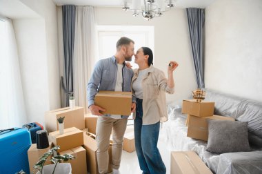 Genç çift yeni evlerine taşınmayı kutluyorlar, ellerinde evin anahtarları ve karton kutular parlak bir odada.