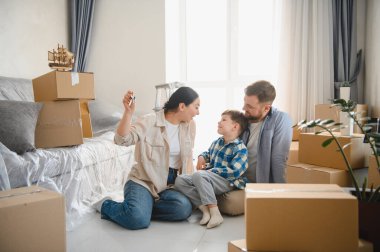 Mutlu bir aile, yeni bir ev satın alıp, elinde anahtarlar, yerde oturup, etrafı karton kutularla dolu boş bir odada kutluyor.