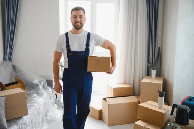 Profesyonel nakliyeci gülümsüyor, paketlenmiş eşyalarla dolu bir odada karton bir kutu tutuyor, ev taşıma ve teslimatı temsil ediyor.