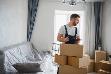 Üniformalı bir adam, karton kutularla dolu bir odada duruyor, yeni bir ev taşınması için tabletteki envanter listesini kontrol ediyor.