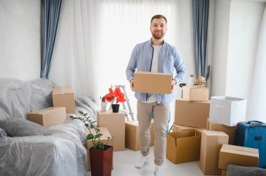 Elinde karton bir kutu olan gülümseyen adam, paketlenmiş kutularla bir odada duruyor, evin yerini değiştirmeyi ve taşınmayı simgeliyor.