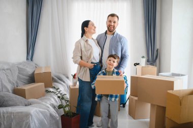 Karton kutularla çevrili yeni evdeki mutlu aile portresi taşınma gününü ve mülkiyet hakkını kutluyor.