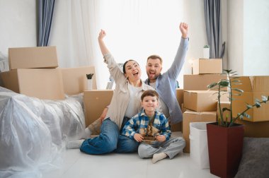 Mutlu bir aile yeni bir eve taşınıyor, karton kutularla çevrili yerde oturuyor, yeni başlangıçları kutluyorlar.